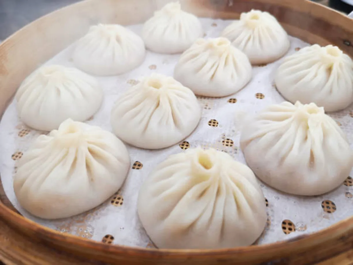 Fresh steaming soup dumplings with pleated gathered tops, arranged inside bamboo steamer on perforated white paper liner, rising soft steam visible.
