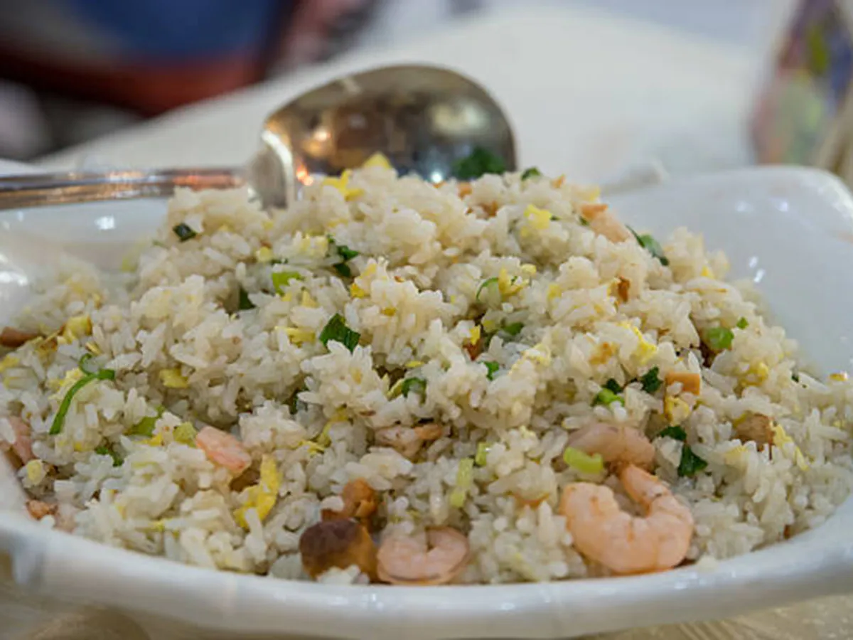 Plateful of seafood fried rice with plump shrimp, scrambled egg bits, green scallions and diced mushrooms, served in square white bowl with metal spoon behind.