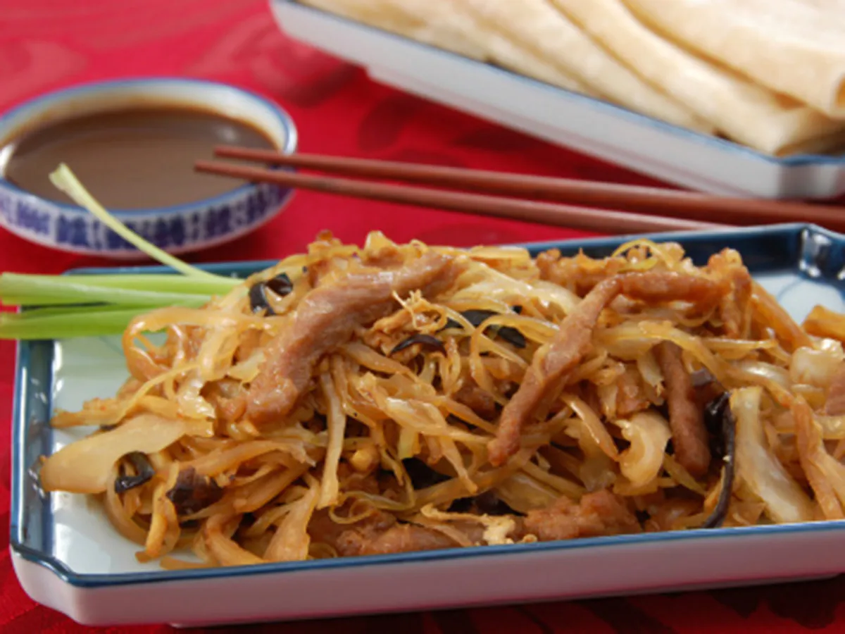 Stir‑fried shredded pork with cabbage and wood ear mushrooms served on blue‑rim rectangular plate. Green scallions, dipping sauce bowl, chopsticks and thin pancakes sit on bright red patterned tablecloth.