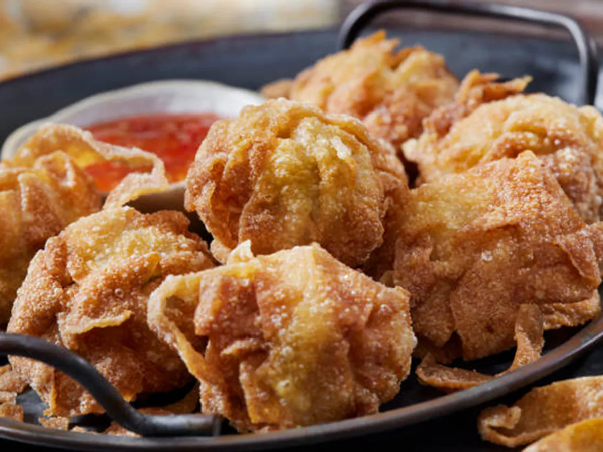 Crispy golden deep‑fried wontons piled on dark metal platter, textured bubbly wrappers, small side bowl of bright red sweet chili dipping sauce.