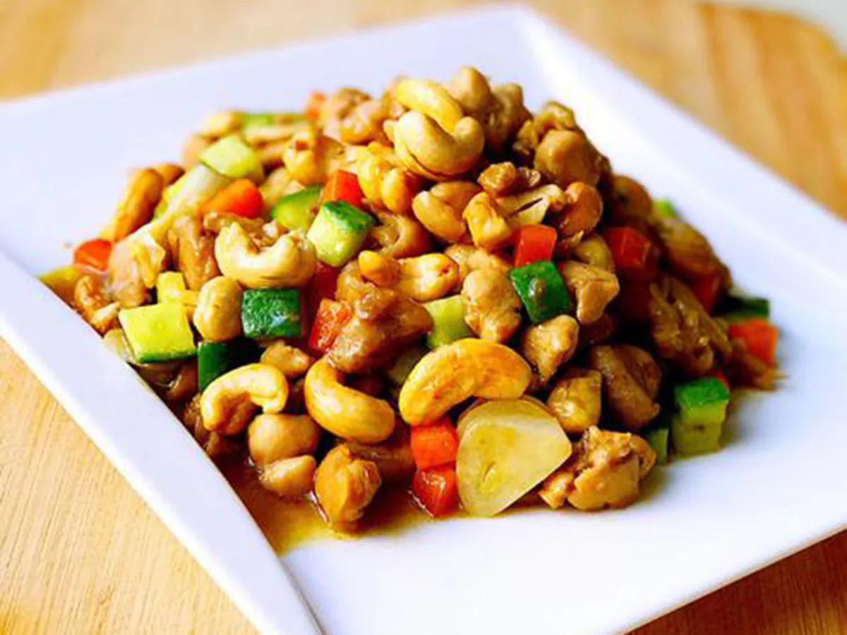 Kung pao chicken stir‑fry on white square plate, tender diced chicken mixed with whole cashew nuts, diced cucumber, red carrot and garlic in glossy savory sauce.