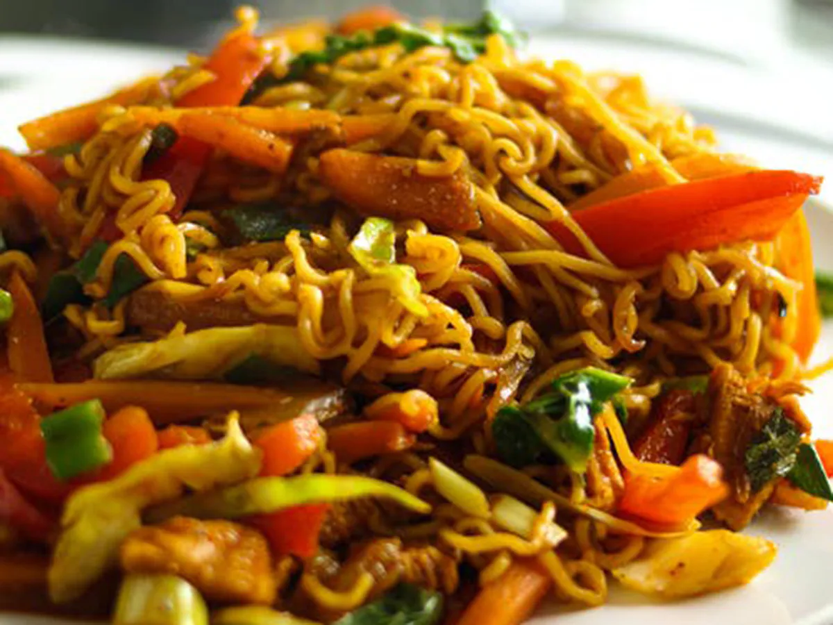 Heaped plate of savory stir‑fried instant noodles tossed with chicken, red bell pepper, carrot, cabbage and leafy greens, coated in rich spiced sauce.